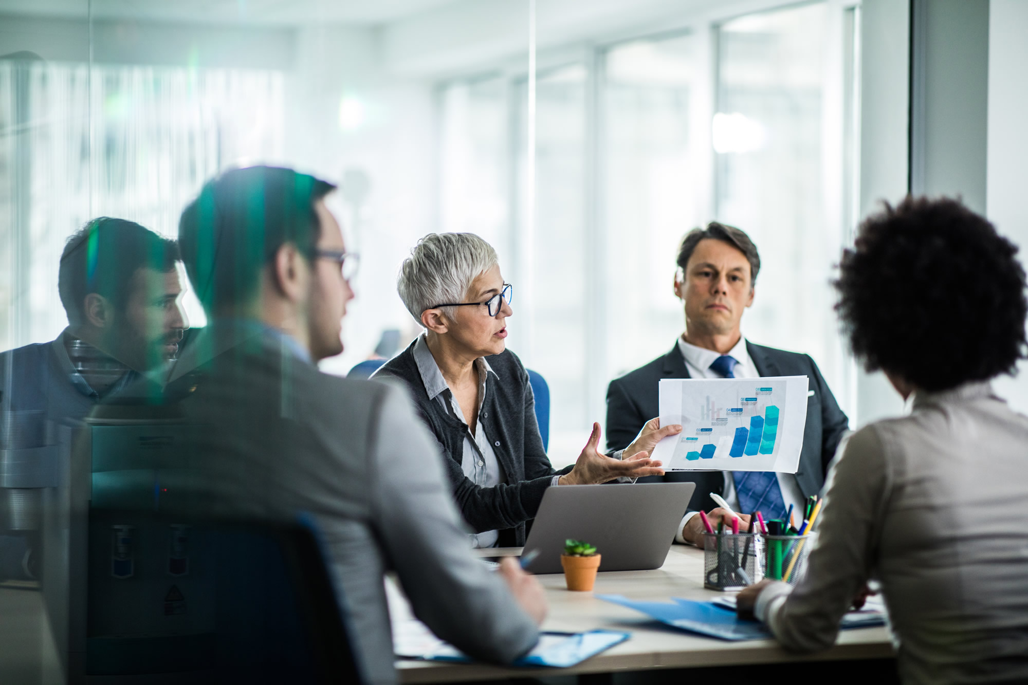 Mature businesswoman talking to her colleagues while examining chart on a meeting in the office. The view is through glass 